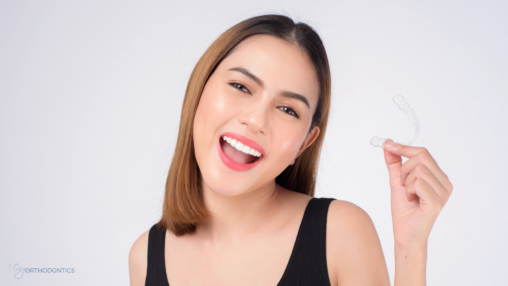 Happy woman holding a clear aligner and smiling, showing off her Invisalign tray during orthodontic care after understanding about Does Insurance Cover Invisalign in Little Falls and Metuchen, NJ